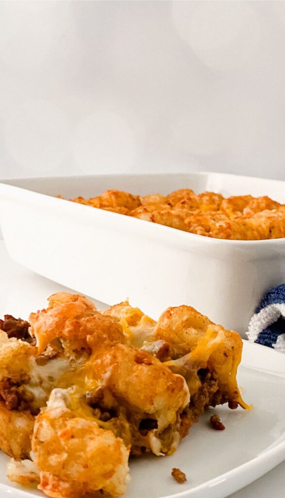 A portion of Tasty Taco Tot Casserole with sour cream on top on a plate in the foreground, with the casserole dish in the background on a white surface.