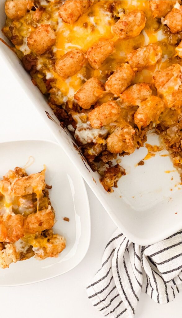 Tasty Taco Tot Casserole: A baked delight with tater tots and melted cheese in a white dish, partially served on a small plate beside a striped cloth.