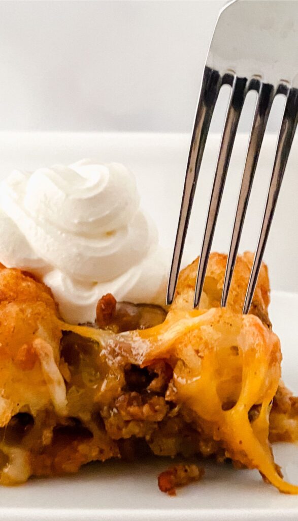 A fork pierces a piece of Tasty Taco Tot Casserole, topped with a dollop of whipped cream on a white plate.