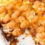 A Taco Tot Casserole dish brimming with ground meat, melted cheese, and a topping of golden-brown tater tots sits invitingly, a tempting portion already missing.