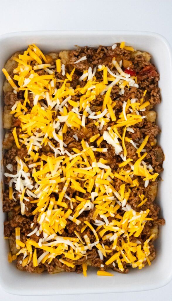 A casserole dish brimming with cooked ground meat and half of the shredded cheese to make the Taco Tot Casserole.