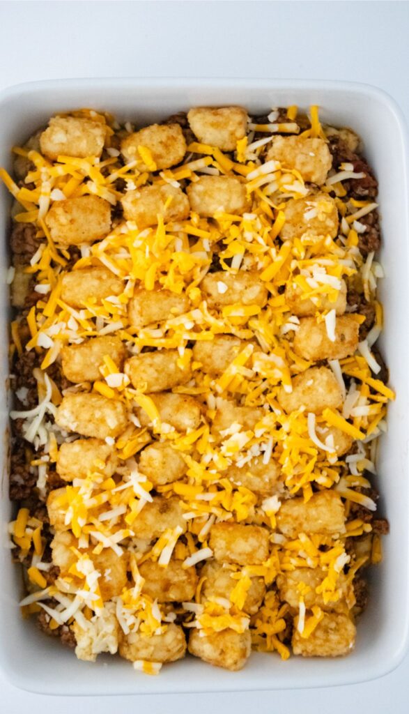 A baking dish with one layer of tator tots, topped with the ground beef layer, tots, and the rest of the cheese before baking a Taco Tot Casserole.