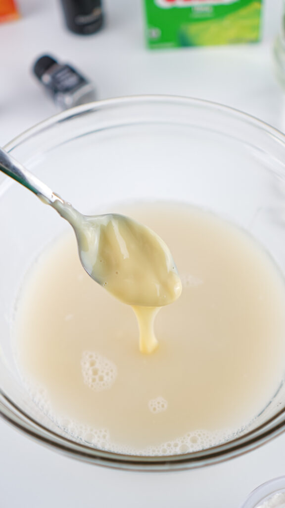 Eagles Jello Shots 6 A spoon drips creamy yellow liquid into a glass bowl filled with off-white Jello shots on a white surface. Various bottles are blurred in the background.