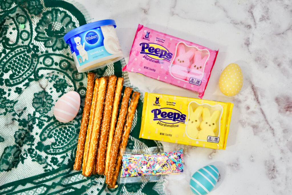 Vanilla frosting, pretzel rods, multicolor sprinkles, yellow and pink Peeps marshmallows, and decorated eggs rest on a green lace cloth atop a marble surface.
