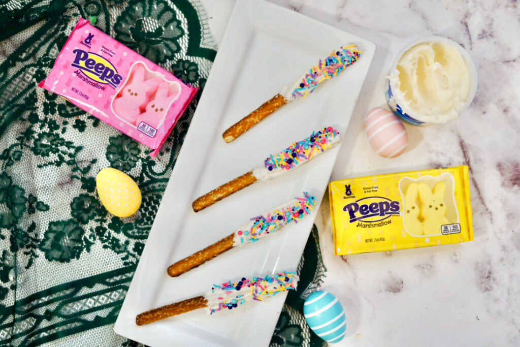 Four decorated Peeps Pretzel Rods with white chocolate and vibrant sprinkles rest on a rectangular plate. Surrounding them are cheerful pink and yellow Peeps packages, a bowl of frosting, and pastel eggs, creating a delightful Easter-themed display.