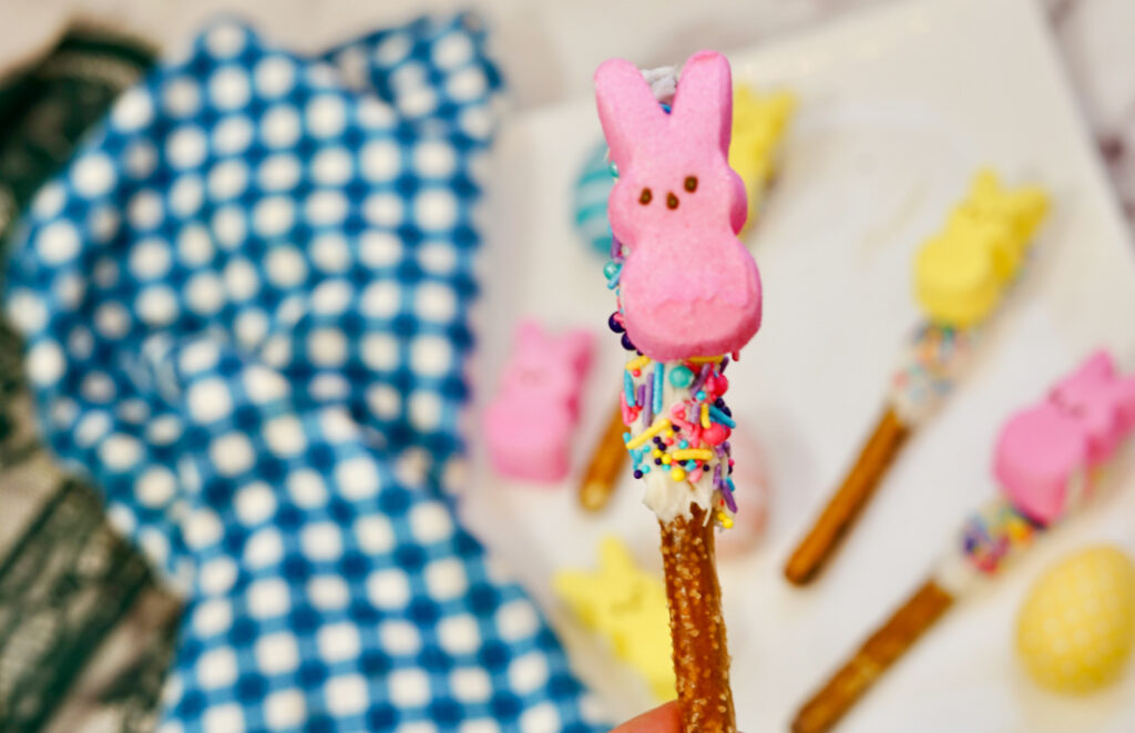 A Peeps Pretzel Rod adorned with white icing, vibrant sprinkles, and a pink marshmallow bunny tops off the sweet treat. Similar decorated pretzels are scattered in the background on a blue and white-checkered cloth.