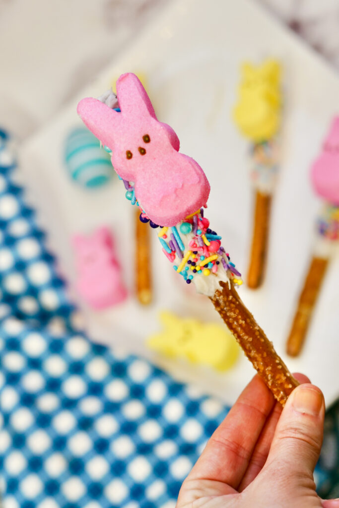 A hand holds a Peeps Pretzel Rod adorned with white chocolate, colorful sprinkles, and topped with a pink marshmallow bunny. In the background, other similarly decorated pretzel rods are arranged on a plate, each a festive treat in its own right.