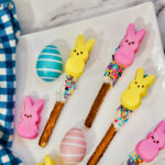 Peeps Pretzel Rods adorned with marshmallow bunnies and sprinkles rest on chocolate-dipped sticks, accompanied by striped and polka-dotted eggs on a white plate with a blue checkered cloth alongside.