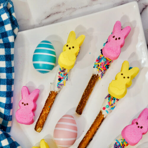 Peeps Pretzel Rods adorned with marshmallow bunnies and sprinkles rest on chocolate-dipped sticks, accompanied by striped and polka-dotted eggs on a white plate with a blue checkered cloth alongside.