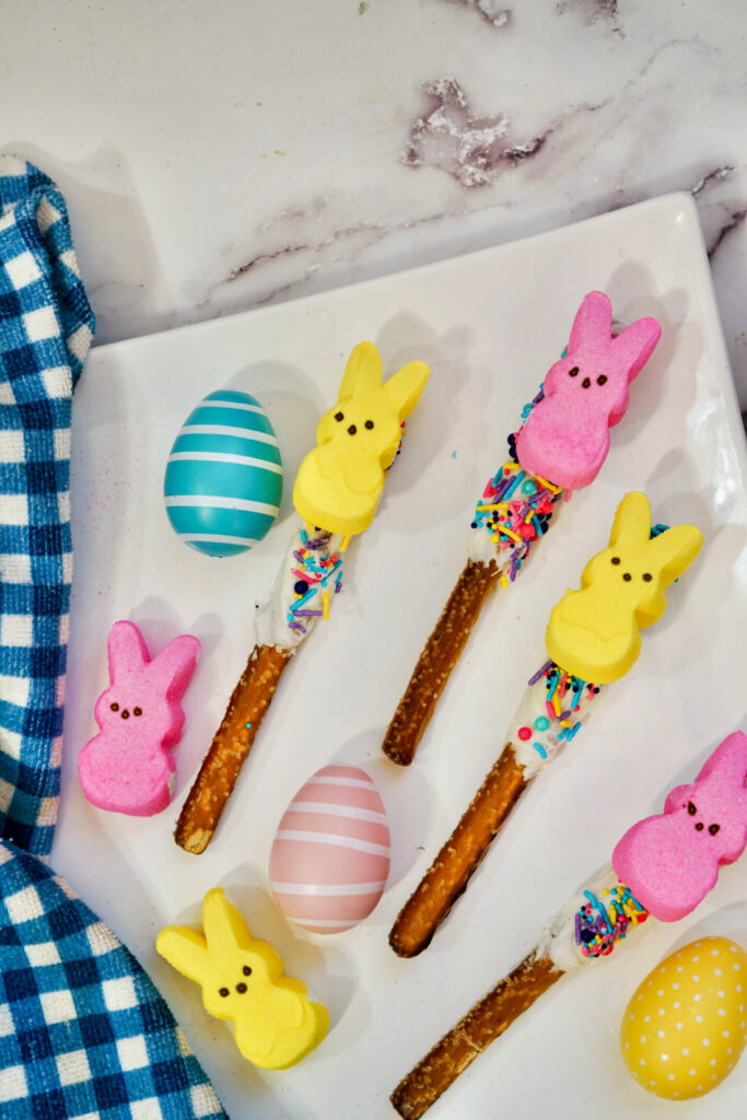 Peeps Pretzel Rods adorned with marshmallow bunnies and sprinkles rest on chocolate-dipped sticks, accompanied by striped and polka-dotted eggs on a white plate with a blue checkered cloth alongside.