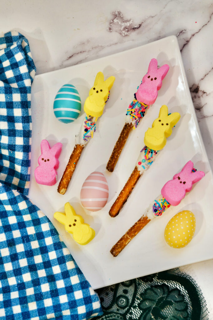 Colorful marshmallow bunnies and pastel eggs on a plate are complemented by Peeps Pretzel Rods, decorated with white chocolate and sprinkles, next to a blue checkered cloth.