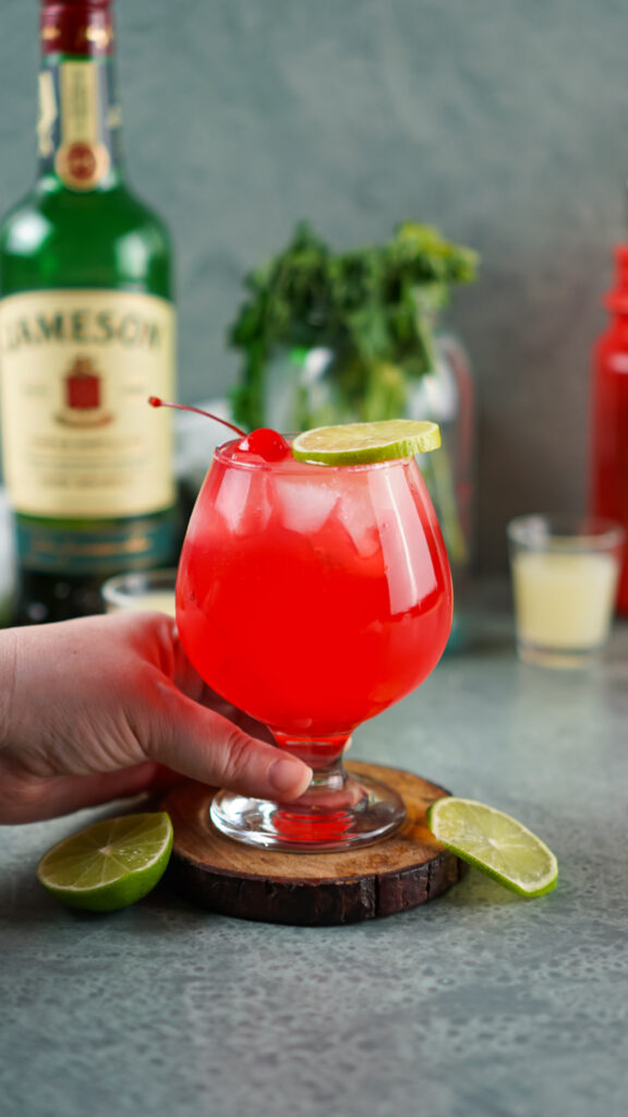 A hand holds an Irish Redhead cocktail in a stemmed glass, adorned with a lime slice and cherry. In the backdrop, a bottle of Jameson is accompanied by fresh lime wedges.