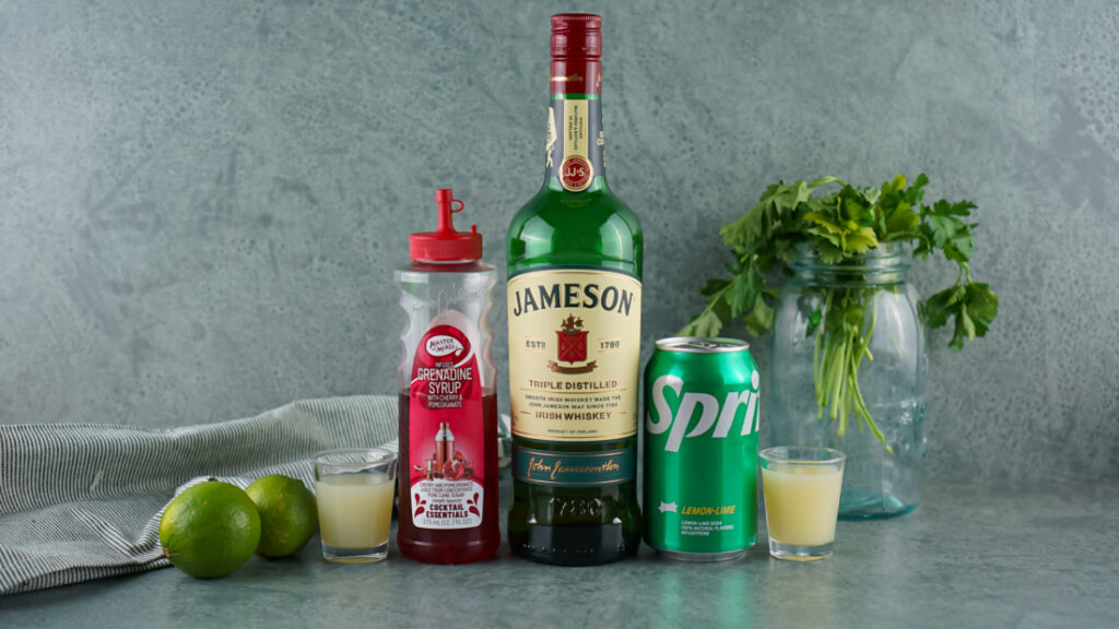 A bottle of Irish whiskey, a can of Sprite, grenadine syrup, and two limes come together to craft the delightful Irish Redhead Cocktail. Beside them, parsley in a jar and a juice-filled glass with a shot glass complete the scene on the gray surface.