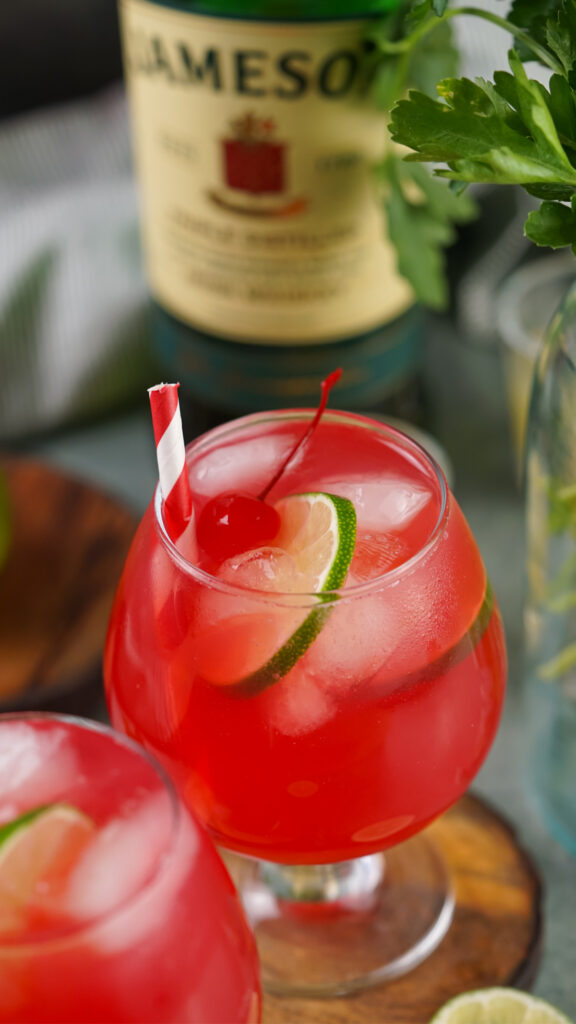 A red cocktail with ice, lime slices, and a cherry, served in a rounded glass with a red and white straw. This delightful Irish Redhead is complemented by a partially visible bottle in the background.