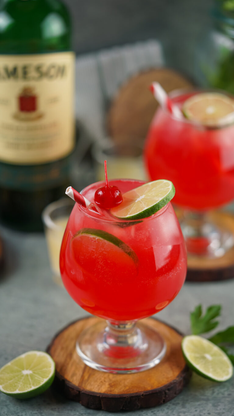 Two Irish Redhead Cocktails are adorned with lime slices and cherries, perched on wooden coasters. In the background, a bottle and extra garnishes complete the inviting scene.