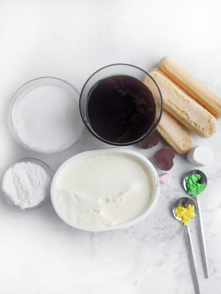 Ingredients for a delightful dessert on a white surface include whipped cream, ladyfingers, coffee, sugar, cocoa powder, marshmallows, and colorful sprinkles in teaspoons. Perfect for crafting an Easter Bunny-inspired Tiramisu with a playful touch of sweetness.