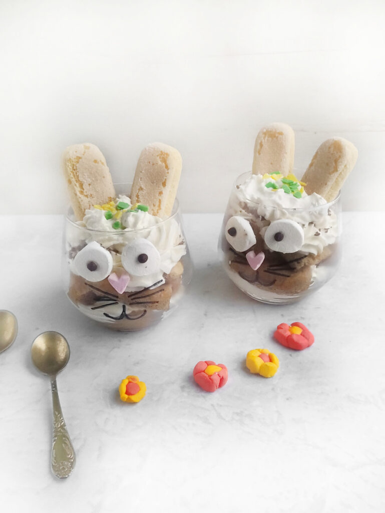 Two bunny-themed desserts in glasses, resembling playful Easter bunnies, come with whipped cream and ladyfingers as ears. They are decorated with candy eyes and flowers, all presented on a marble surface beside two spoons.