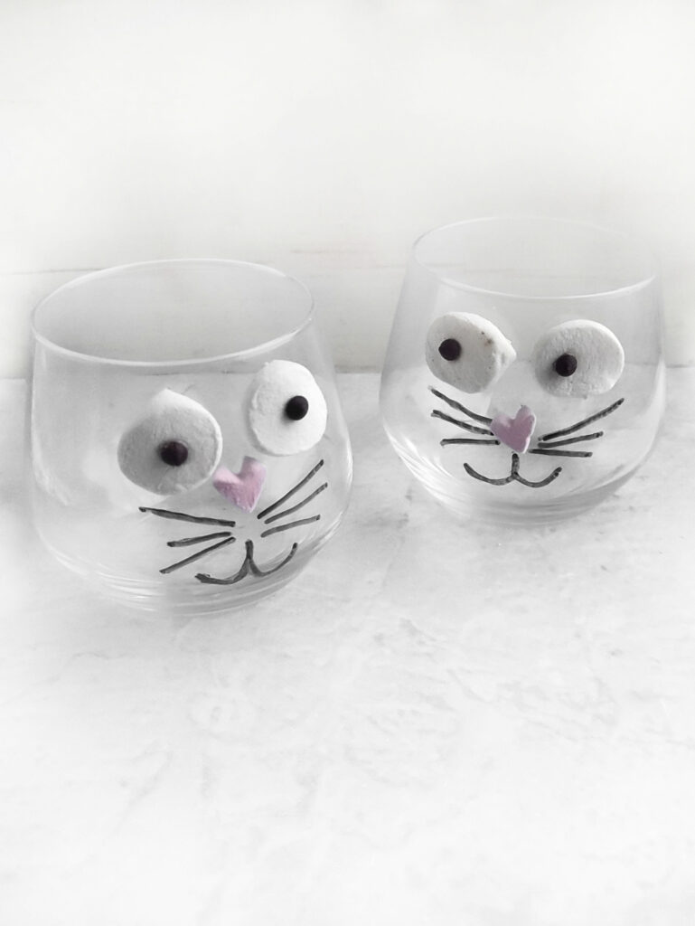 Two clear glasses, decorated with cat faces, bring a whimsical vibe reminiscent of an Easter Bunny. Marshmallows serve as eyes and pink candy forms the noses, resting on a light surface like a sweet rendition of tiramisu.