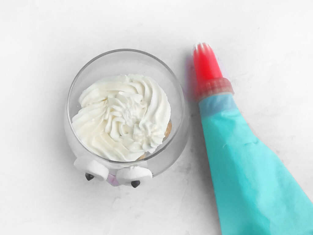A dessert in a clear bowl with googly eyes, topped with swirled whipped cream, creates a whimsical Easter Bunny vibe. A blue piping bag with a red tip rests beside it on a light surface, adding a playful touch reminiscent of crafting tiramisu decorations.