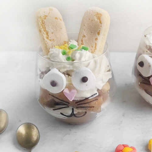 A glass filled with dessert, decorated to resemble an Easter Bunny tiramisu with cookies as ears, marshmallow eyes, and a heart-shaped nose. Two spoons and small candy flowers are nearby.