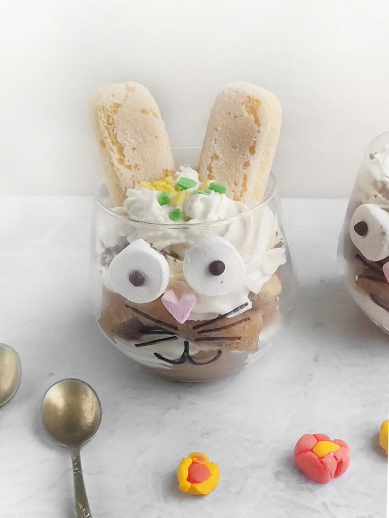 A glass filled with dessert, decorated to resemble an Easter Bunny tiramisu with cookies as ears, marshmallow eyes, and a heart-shaped nose. Two spoons and small candy flowers are nearby.
