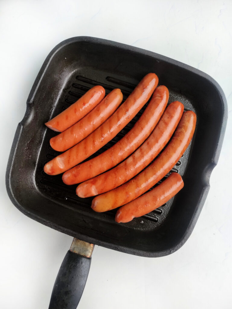 Seven sausages are arranged in a grill pan, ready to be cooked on a white surface—perfect for your next hot dog celebration.