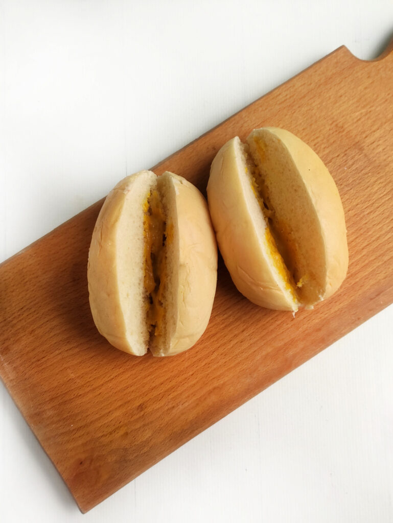 Two plain bread rolls, each sliced in half and filled with a yellow spread, are displayed on a wooden celebration board against a white background.