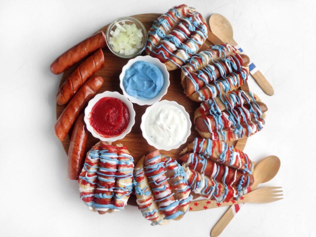 A round Hot Dog Patriot Celebration Board featuring hot dogs with red, white, and blue sauce, sausages, bowls of chopped onions, ketchup, blue and white sauces, plus wooden cutlery and napkins—perfect for a festive gathering.