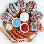 A Hot Dog Patriot Celebration Board: a wooden tray featuring hot dogs drizzled in red, white, and blue sauces, tasty dips in small bowls, chopped onions, three plain sausages, and decorated wooden utensils.
