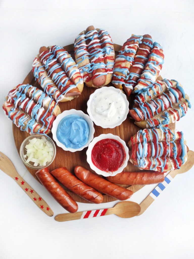 A Hot Dog Patriot Celebration Board: a wooden tray featuring hot dogs drizzled in red, white, and blue sauces, tasty dips in small bowls, chopped onions, three plain sausages, and decorated wooden utensils.