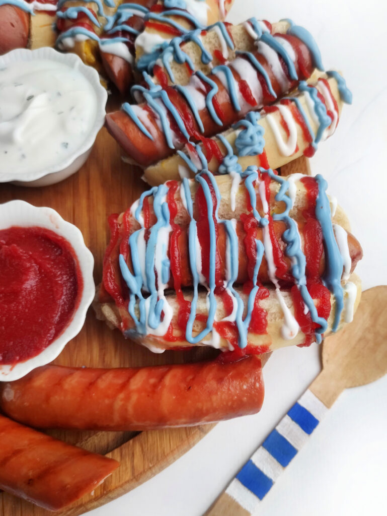 Hot dogs take center stage on a Patriot Celebration Board, topped with ketchup and blue and white sauces. Served with bowls of dipping sauce, sausage pieces, and a wooden fork for the perfect festive bite.