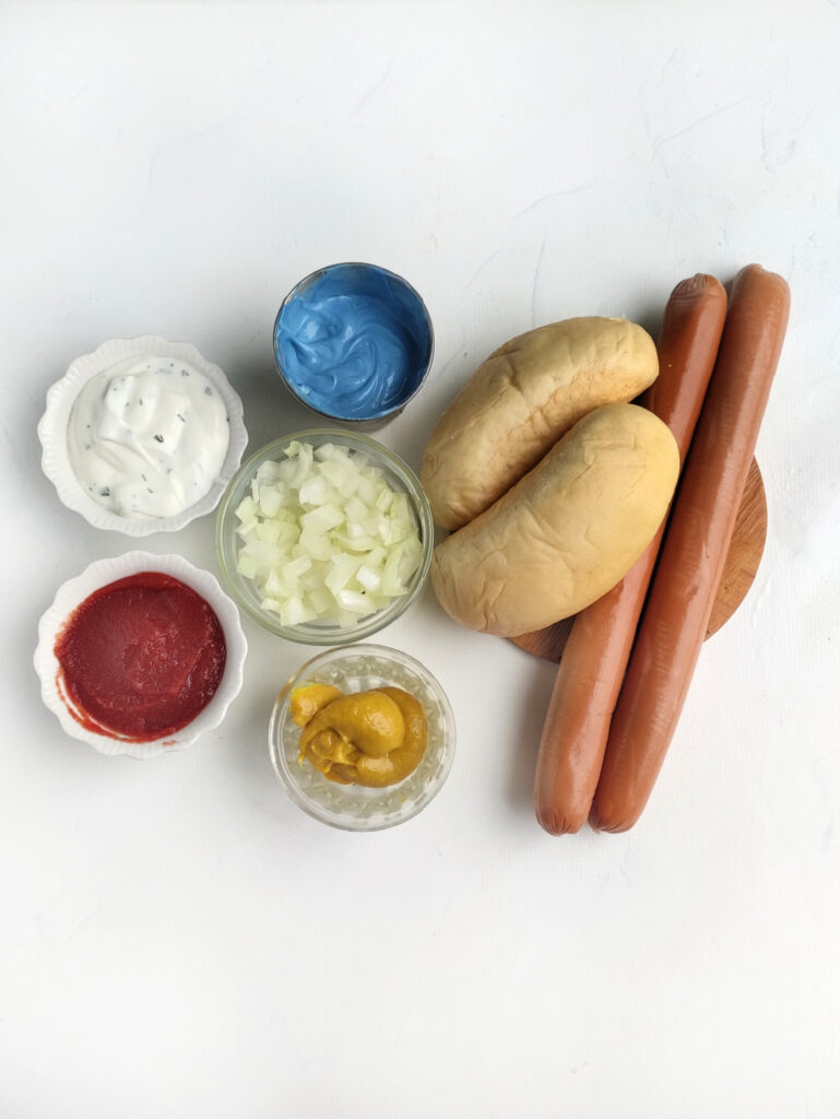 Two hot dog buns and sausages are paired with small bowls of diced onions, ketchup, mustard, blue sauce, and white sauce on a white background—perfect for your Hot Dog Patriot Celebration Board.