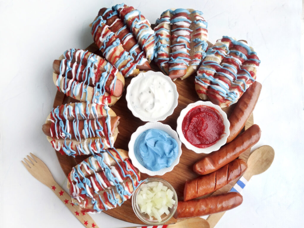 A round Hot Dog Patriot Celebration Board featuring grilled hot dogs topped with festive red, white, and blue sauce, served with ketchup, ranch, blue dip, and chopped onions. Wooden utensils are on the side for easy serving.