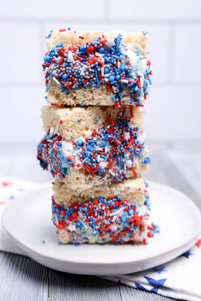 Three No-Bake Patriotic rice crispy treats, topped with red, white, and blue sprinkles, are stacked on a white plate against a light background.
