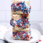 Three No-Bake Patriotic Rice Krispies treats stacked on a white plate, decorated with red, white, and blue sprinkles and icing, with a star-patterned napkin in the background.