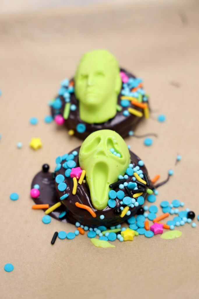 Two Summerween Slasher Oreos topped with green candy faces, colorful sprinkles, and star-shaped decorations rest on a brown surface.