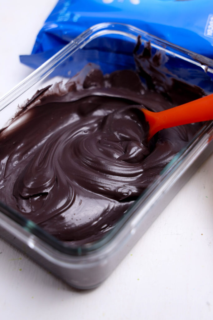 A glass dish filled with smooth, melted dark chocolate being stirred with an orange spatula—perfect for dipping Summerween Slasher Oreos or your favorite spooky Summerween treats.