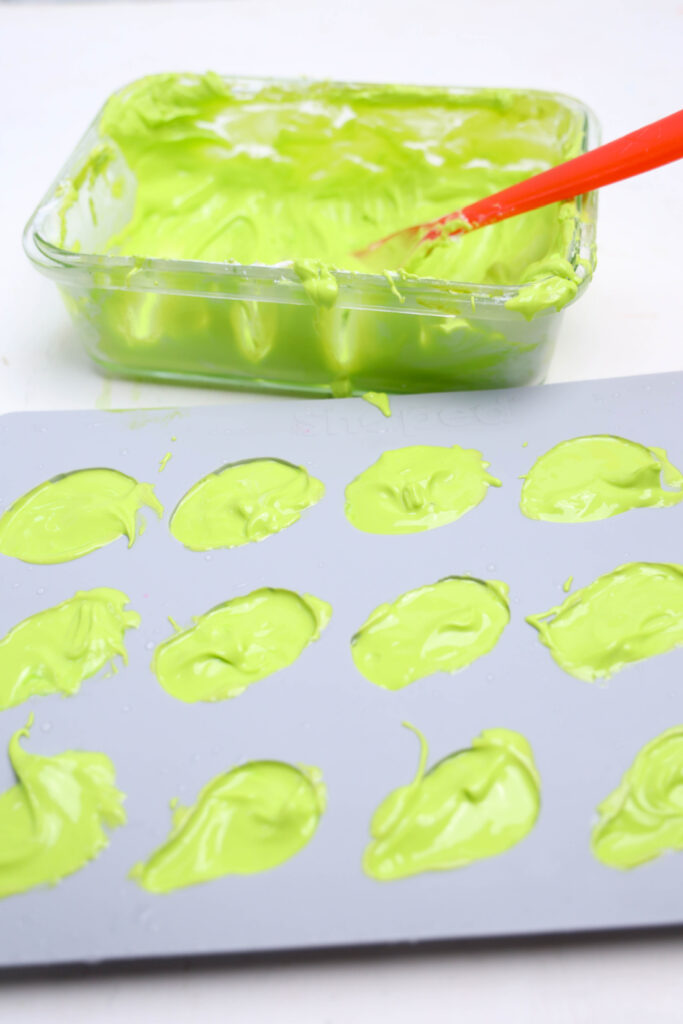 A silicone mold filled with green creamy mixture sits in front of a glass dish containing more of the mixture, a red spatula, and crushed Oreos—perfect for your next Summerween treat.