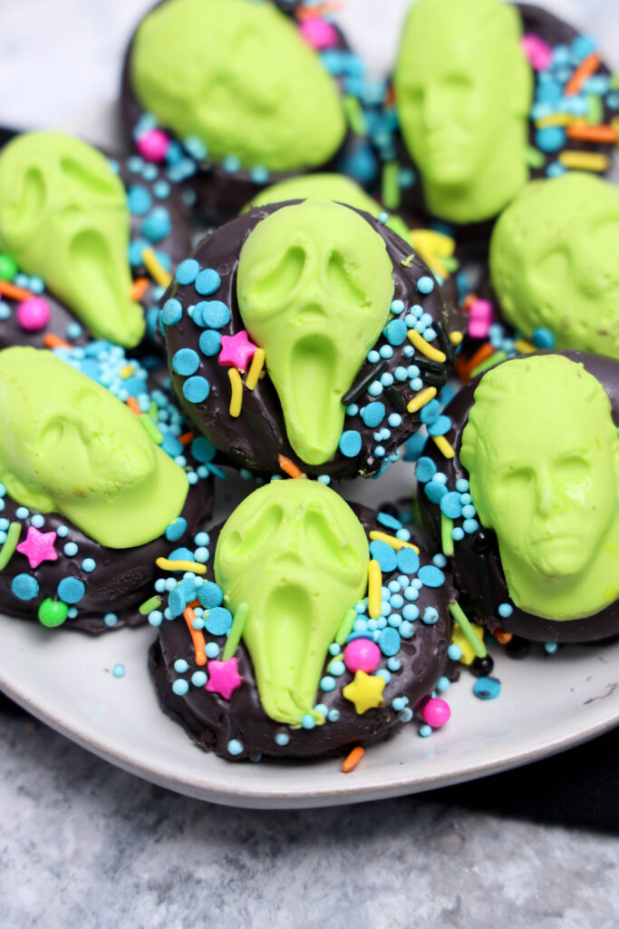 Chocolate-covered cookies, called Summerween Slasher Oreos, are topped with scream-faced and skull-shaped candies, plus colorful sprinkles, served on a white plate—perfect for a spooky Summerween treat!.