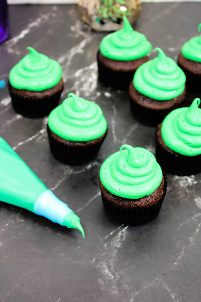 Spooky Halloween Eyeball Cupcakes: The Perfect Creepy-Cute Treats 4 Chocolate cupcakes with green frosting are arranged on a dark marble surface, with a piping bag filled with green icing placed beside them, perfect for creating spooky Halloween Eyeball Cupcakes.