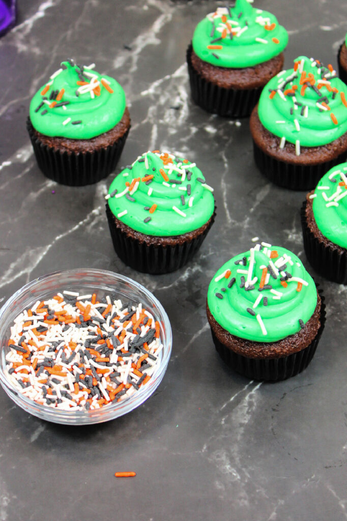 Spooky Halloween Eyeball Cupcakes: The Perfect Creepy-Cute Treats 5 Spooky Halloween Eyeball Cupcakes with green frosting and orange, white, and black sprinkles are arranged on a dark marble surface next to a small bowl of sprinkles.