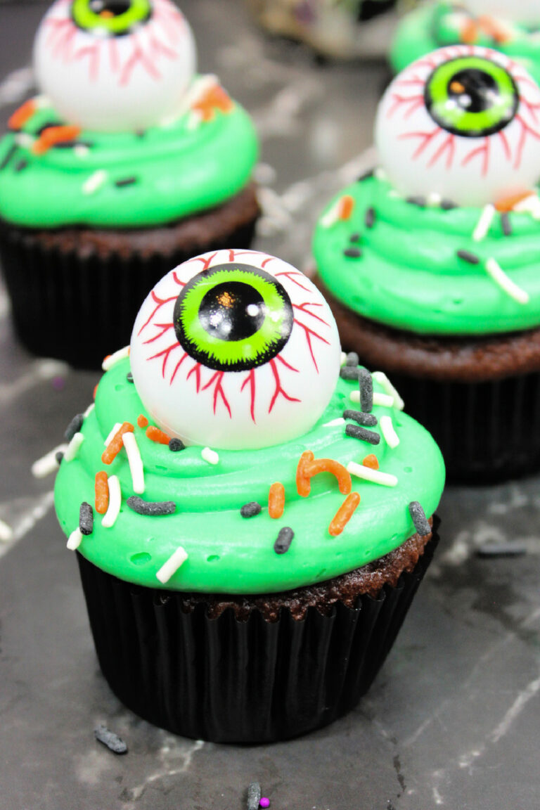 Chocolate cupcake with bright green frosting, colorful sprinkles, and a large, realistic candy eyeball on top—perfect for Spooky Halloween Eyeball Cupcakes displayed on a dark surface.