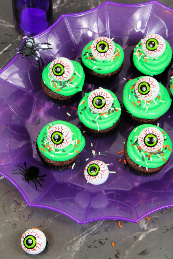 Spooky Halloween Eyeball Cupcakes: The Perfect Creepy-Cute Treats 9 Spooky Halloween Eyeball Cupcakes with green frosting and eyeball decorations are arranged on a purple spiderweb platter, surrounded by plastic spiders and fake eyeballs on a gray surface.