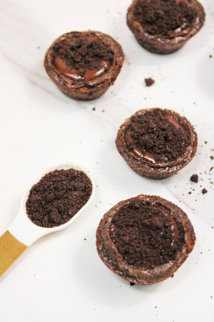 Four chocolate tarts topped with chocolate crumbs are arranged on a white surface, resembling mini Halloween brownies, with a spoonful of chocolate crumbs beside them.