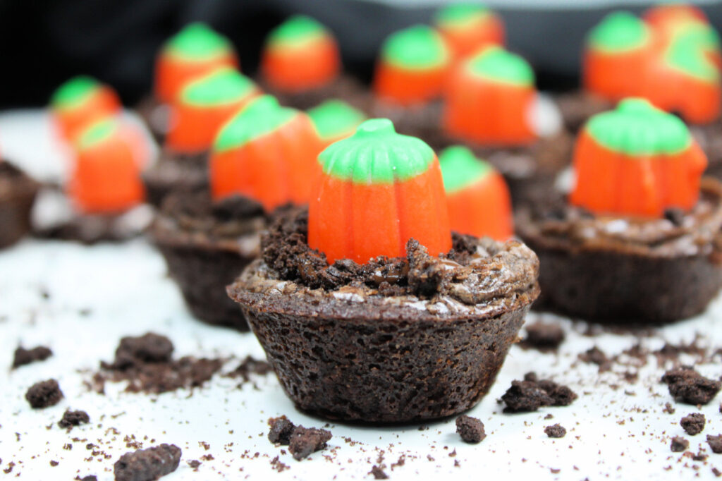 Mini Halloween brownies are topped with crushed cookies and candy pumpkins, creating adorable pumpkin patch brownies arranged on a white surface with cookie crumbs scattered around.
