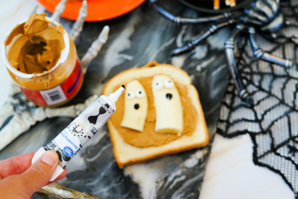 Halloween Toast That'll Make You Go BOO-nanas 7 A slice of Halloween Toast with peanut butter is topped with two banana ghosts decorated with icing. In the foreground, a hand holds a tube of black decorating gel, perfect for spooky finishing touches.