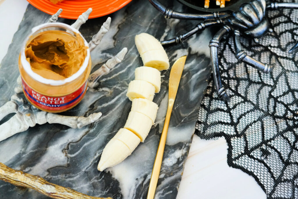 Halloween Toast That'll Make You Go BOO-nanas 5 A sliced banana on a marble surface with a gold knife, an open jar of peanut butter, plastic skeleton hands, a fake spider, and black lace web decor—perfect for crafting spooky Halloween Toast.