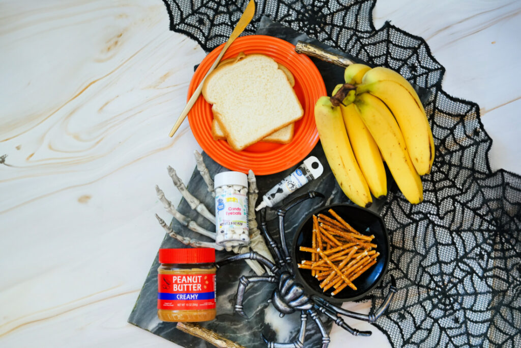 Halloween Toast That'll Make You Go BOO-nanas 3 A Halloween Toast setup featuring bread, peanut butter, bananas, candy eyes, pretzel sticks, and skeleton hand props arranged on a web-patterned cloth for a spooky snack display.