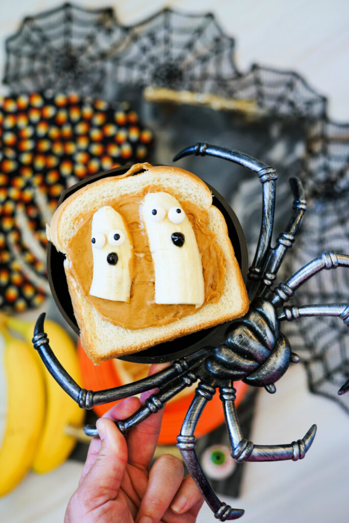 Halloween Toast That'll Make You Go BOO-nanas 8 A hand holds a slice of Halloween Toast with peanut butter and two banana slices decorated as ghosts. A large fake spider and spooky, Halloween-themed decorations fill the background.