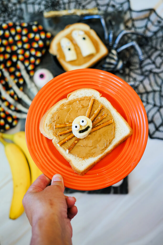 Halloween Toast That'll Make You Go BOO-nanas 9 A hand holds an orange plate with a slice of Halloween Toast—bread topped with peanut butter, pretzel sticks, and a candy eye arranged like a spider. Halloween-themed food items are visible in the background.